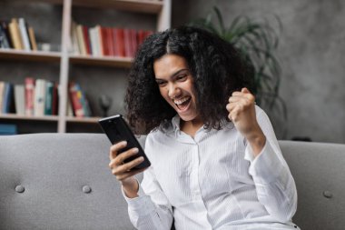 Rahat koltukta oturan, modern akıllı telefonlu, gündelik giysili çekici genç bir kadın. Evde boş zamanlarında internette sörf yapan gülümseyen bir kadın..