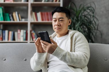 Gündelik giyimli çekici Asyalı adam rahat koltukta oturuyor ve elinde modern akıllı telefon var. Evde boş zamanlarında internette sörf yapan gülümseyen bir erkek..