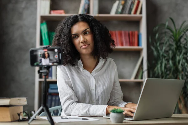 stock image Pleasant young woman working on laptop while recording video on her smart phone on tripod. Multinational female freelancer working remotely while staying at home.