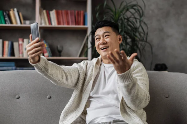 現代的なスマートフォンを手に快適なソファに座ってカジュアルな服装で魅力的なアジア人男性 自宅で余暇の間にビデオ通話をしている男性の笑顔 — ストック写真