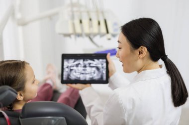 Gülümseyen mutlu beyaz çocuk dişçiyi ziyaret ederken, modern ışık hastanesi kliniğinde dişçi koltuğunda otururken. Genç Asyalı dişçi elinde röntgen taramasıyla tablet tutuyormuş..