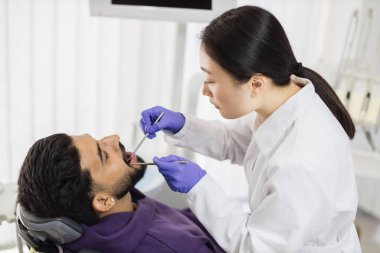 Beyaz üniformalı, eldivenli, yakışıklı Asyalı genç bir doktor erkek hastasına bakıcılık yapıyor ya da diş ağrısı tedavisi uyguluyor. Sakallı, dişçi koltuğunda oturan çekici bir adam..