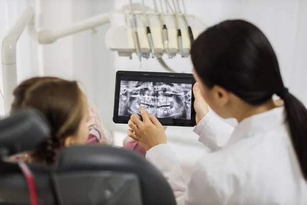Back view of smiling happy caucasian child girl visiting dentist, sitting in dental chair at modern light hospital clinic. Young asian woman dentist holding tablet pc with x ray scan.