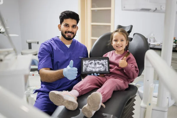 Pretty smiling preschool girl, sitting on dentist chair and looking at digital tablet with x-ray scan image of teeth together with her professional male bearded dentist at clinic showing thumbs up.