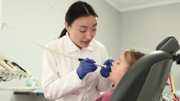 开枪射击身穿白衣的年轻漂亮的亚裔女牙医 为她可爱的小女孩在诊所的牙椅上为她做牙齿检查和龋病治疗 — 图库视频影像
