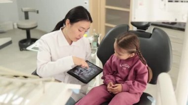 Gülümseyen mutlu beyaz kız dişçiyi ziyaret ediyor, modern ışık hastanesi kliniğinde dişçi koltuğunda oturuyor. Genç Asyalı dişçi elinde röntgen taramasıyla tablet tutuyormuş..