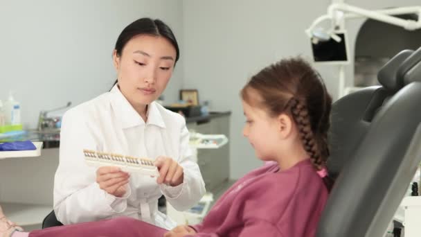 年轻快乐的学龄前小女孩穿着休闲装在牙科诊所里 轻松而现代的办公室里 身穿白衣的亚洲女医生将病人的牙齿与牙齿美白图进行比较 — 图库视频影像