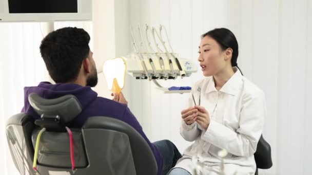 在新的明亮的牙科诊所里 漂亮的亚洲女牙医笑着看病人时 英俊的大胡子男人一边检查牙齿 一边照镜子 这是他的肖像 — 图库视频影像