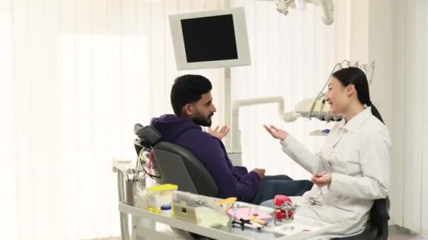 在牙科医院接受牙齿治疗 满脸笑容的满脸胡须的男人坐在牙医椅子上 与兴奋的美丽的亚裔牙医女人谈论着现代牙科 — 图库视频影像