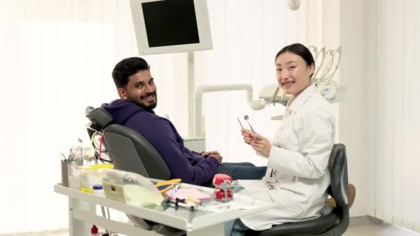 在牙科医院接受牙齿治疗 在轻盈的现代牙科诊所里 满脸笑容的满脸胡须的男人坐在牙医椅子上 与兴奋的美丽的亚裔女牙医坐在一起 看着相机 — 图库视频影像
