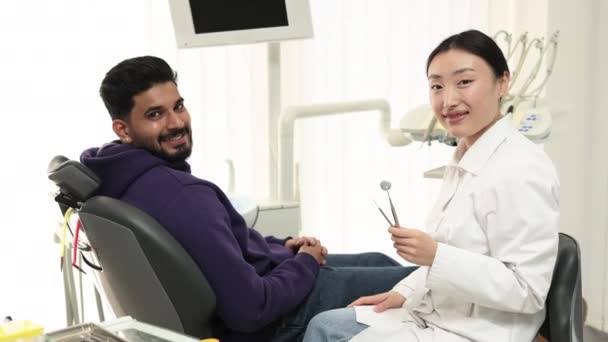 歯科病院で歯の治療 笑顔ひげを生やした男に座っています歯科椅子で軽い現代の歯科クリニック 親指を示していますまで興奮美しいアジアの女性歯科医とカメラを見て — ストック動画
