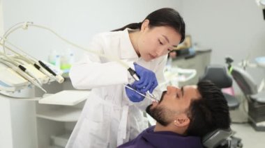 Mutlu hasta ve dişçi konsepti. Genç, güzel Asyalı kadın stomatolog diş matkabı kullanarak yakışıklı, sakallı bir gencin dişlerini tedavi ediyor. Ağız sağlığı ve hijyen.