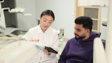 Tıp, dişçilik ve oral bakım konsepti. Asyalı dişçi endişeli erkek hastaya tablet bilgisayar gösteriyor. Modern diş kliniğinde sakallı genç bir adam..