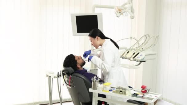 美丽的笑与白色的牙齿 亚洲牙医通过牙科诊所的放大镜 用牙钻治疗一个年轻大胡子男子的牙齿 — 图库视频影像