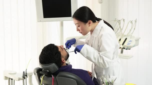 美丽的笑与白色的牙齿 亚洲牙医通过牙科诊所的放大镜 用牙钻治疗一个年轻大胡子男子的牙齿 — 图库视频影像