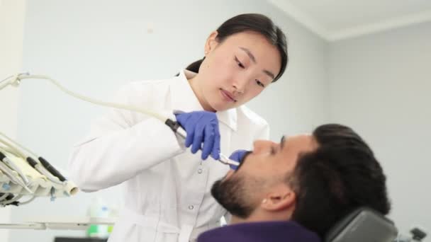 歯科医院現代事務所で歯科処置中に髭を生やしたハンサムな男性 若い美しいアジアの女性歯科医は 歯科ドリルとミラーを使用して 男の歯を硬化 — ストック動画
