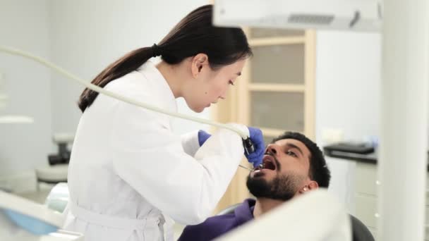 开枪射击身穿白衣的年轻漂亮的亚裔女牙医 为她漂亮的男性病人做牙齿检查和龋病治疗 她的胡子男子坐在诊所的牙椅上 — 图库视频影像