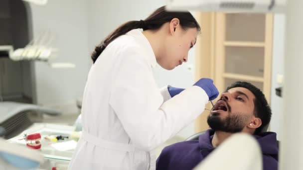 身穿白衣戴手套的年轻漂亮的亚洲医生的侧视图 为她的男性病人做检查或治疗龋和牙痛 漂亮的胡子男子坐在牙椅上 — 图库视频影像
