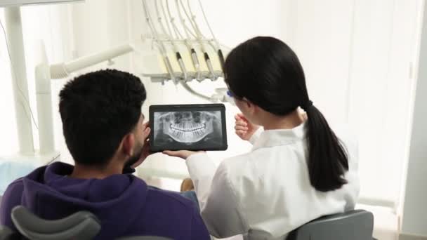 若い魅力的な男が歯科医を訪問し 現代のライト病院の診療所で歯科椅子に座ってのバックビュー 若いアジアの女性歯科医は タブレットを保持X線画像 写真を中心に — ストック動画
