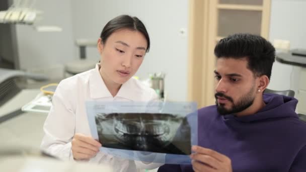 迷人的年轻留着胡子的男人 坐在牙医椅子上 和他快乐的亚裔女牙医一起在诊所看X光的牙齿扫描照片 — 图库视频影像