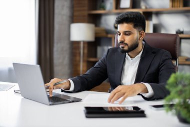 Takım elbise giyen kendine güvenen işadamı notlar ya da mali rapor yazıyor, dizüstü bilgisayarla masada oturuyor, kağıt belgelerle çalışan ciddi bir adama odaklanıyor, öğrenci çevrimiçi çalışıyor, araştırma yapıyor.