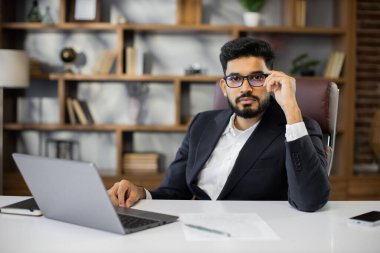 Ofiste çalışırken bilgisayarını kullanan sakallı genç bir iş adamı. Ahşap masada oturan, resmi takım elbiseli, konsantre olmuş, başarılı bir adam..