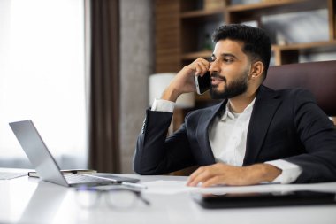 Sandalyede oturan genç sakallı adamın portresi modern ofisteki iş ortaklarıyla telefon görüşmesi yapıyor. Masada, evde, dizüstü bilgisayarla çalışan genç bir işadamı..