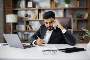 Başarılı genç girişimci dikkatli ve yoğun bakışlı telefon görüşmesi yaparken, modern ofiste masa başında otururken.