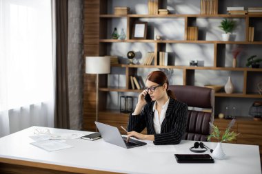 Resmi çağrı. Gözlüklü, kendine güvenen genç ve güzel bir kadın iş ortaklarını arıyor, yeni anlaşma hakkında konuşuyor ve soruyor, modern ofiste dizüstü bilgisayarla çalışıyor..