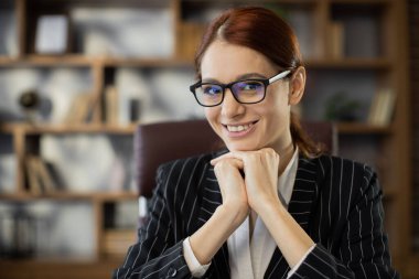 Ofiste oturmuş kameraya bakan, resmi giyinmiş ve gözlüklü neşeli, kızıl saçlı bir kadının yakın plan yüzü. Kızıl saçlı güzel bayan uzaktan kumandalı çalışıyor ya da çalışıyor..