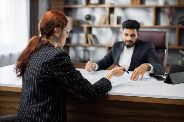 Привлекательный бородатый бизнесмен, сидящий за столом, показывает несколько документов своей коллеге в очках напротив него в современном офисе. Селективный акцент на деловой леди.