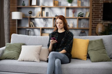 Çekici beyaz saçlı kadın kanepede oturuyor ve modern akıllı telefonu kapalı alanda kullanıyor. Evde çalışma sürecinde ara veren serbest çalışan kadın..