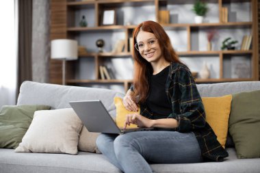 İşi için araştırma yapan genç bir kadın. Gülümseyen kadın kanepeye oturmuş online alışveriş sitesine göz gezdiriyordu. Evde boş zamanlarında internette gezinen mutlu kız..