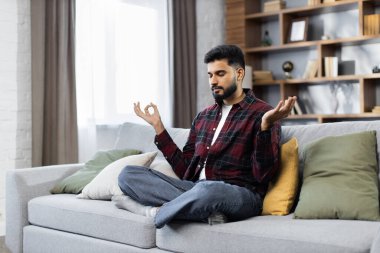 Yong erkeği nilüfer çiçeğiyle kanepede oturup ellerini kucağına koyuyor parmaklarını kavuşturup gözlerini kapatıyor meditasyonun tadını çıkarıyor evde yoga egzersizi yapıyor. Stres yok. Sağlıklı alışkanlık. Farkındalık.