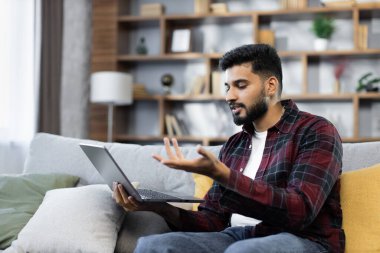 Kırmızı tişörtlü genç sakallı iş adamı dizüstü bilgisayarla çalışıyor, evdeki rahat koltukta otururken internette konuşuyor. Evden uzak çalışma kavramı.