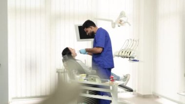 Beyaz üniformalı ve eldivenli sakallı bir erkek doktorun kadın hastası için dişlerini ve dişlerini muayene edişinin yan görüntüsü. Diş sandalyesinde oturan çekici, genç Asyalı kadın..