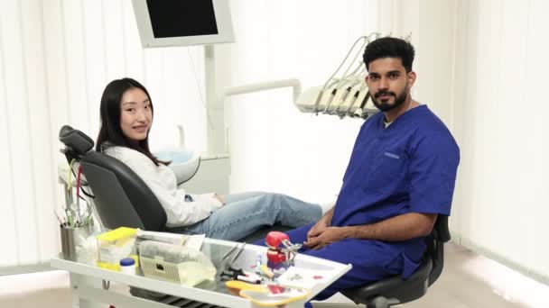 在牙科医院接受牙齿治疗 在轻盈的现代牙科诊所里 坐在牙医椅子上的亚洲女人笑着 带着工具的年轻牙医 看着露出大拇指的相机 — 图库视频影像