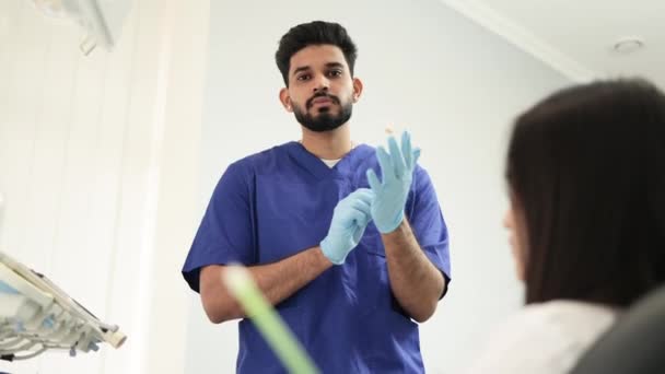 满脸胡须的男牙医头戴蓝色橡胶手套 站在牙科诊所 准备检查和治疗他的女病人 快乐病人和牙医的概念 — 图库视频影像