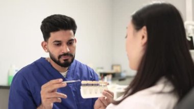 Diş tonu paleti. Kendine güvenen sakallı erkek kozmetik uzmanı dişçi klinikte beyazlatmak için çekici Asyalı kadın dişlerinin rengini seçiyor..