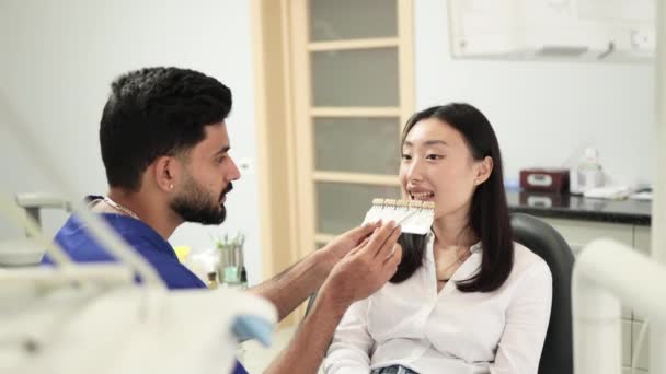 パレットから充填の色を選択歯科医 男性歯科医のバックビュー現代病院で室内で若いアジアの女性の歯の色をチェックし — ストック動画