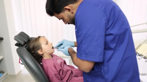 現代の小児歯科医院の歯科処置中にかわいい女の子 子供の魅力的な男性ひげそり歯科医は 歯科ドリルとミラーを使用して 女の子の歯を硬化 — ストック動画