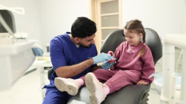 Kendine güvenen genç sakallı pediatri dişçisinin, anaokulundaki küçük kıza, modern diş muayenehanesinde takma dişlerini nasıl fırçalayacağını göstermesi. Pediatrik dişçilik.