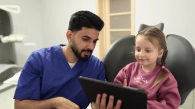 Tıp, pediatrik dişçilik ve oral bakım konsepti. Kendine güvenen erkek, gülümseyen profesyonel dişçi mutlu çocuk hastaya tablet bilgisayar gösteriyor, modern diş kliniğindeki beyaz anaokulu kızı.
