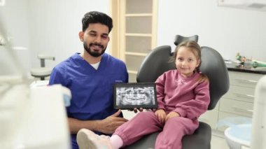 Gülümseyen Kafkasyalı anaokulu kızı, dişçi koltuğunda oturuyor ve dişlerinin röntgen taramasıyla dijital tablete bakıyor. Klinikteki erkek sakallı dişçisiyle birlikte..