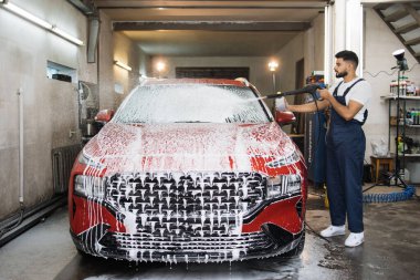 Genç hoş oto yıkama işçisinin portresi, koruyucu tulumlar giymesi, araba yıkamada yüksek basınçlı su jeti ile otomobili temizlemesi, temizlik köpüğünü püskürtmesi. Araba yıkama..