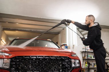 Yüksek basınçlı su kullanarak arabayı temizlemek. Yakışıklı genç adam araba yıkama servisinde yüksek basınçlı su altında modern kırmızı arabayı yıkıyor..