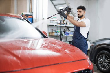 Yüksek basınçlı su kullanarak arabayı temizlemek. Yakışıklı genç adam araba yıkama servisinde yüksek basınçlı su altında modern kırmızı araba kaputunu yıkıyor..