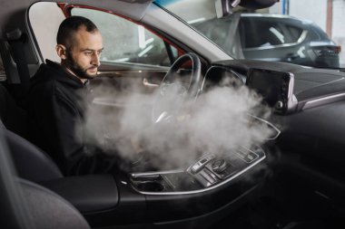 Oto temizleme ve detay verme servisi. Üniformalı yakışıklı sakallı adam arabanın içini sıcak buhar temizleyicisiyle temizliyor. Erkek yüzüne odaklan..