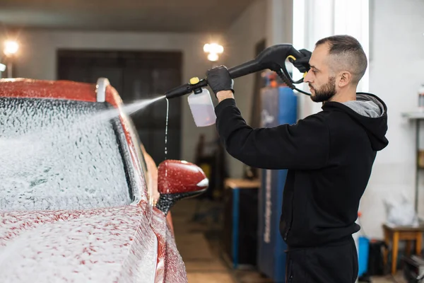 Bearded young man car wash worker spraying cleaning foam to a modern red car holding a high pressure washer. Modern car and foam washing.
