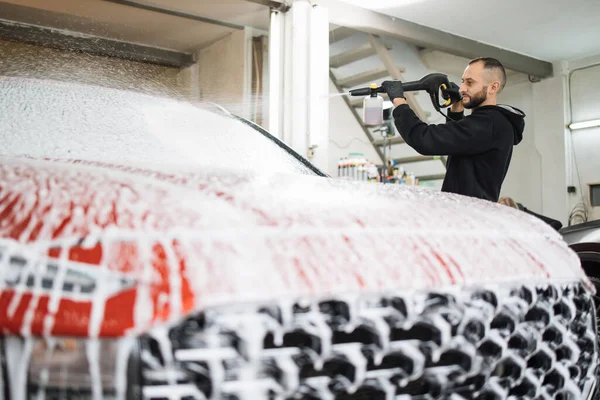 Bearded young man car wash worker spraying cleaning foam to a modern red car holding a high pressure washer. Modern car and foam washing.